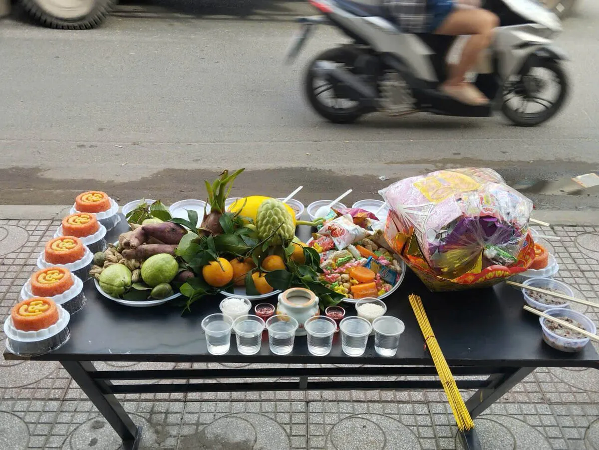 Mâm cúng cô hồn chay gói tiết kiệm bày trên bàn đen ngoài vỉa hè gồm 6 phần xôi gấc chữ Lộc, cháo trắng, chè đậu, đĩa khoai lang đậu phộng ổi, mâm ngũ quả bưởi mãng cầu cam và túi quần áo chúng sinh