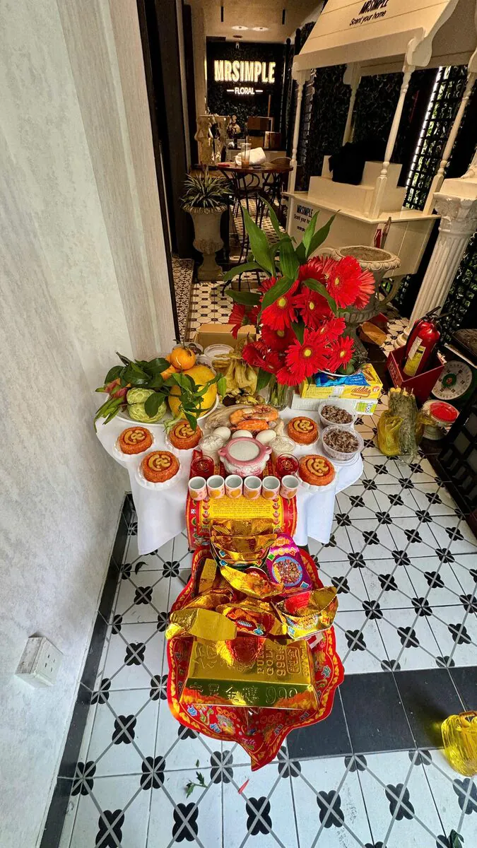Mâm cúng gói tiêu chuẩn đặt tại sảnh trước cửa hàng MrSimple Floral gồm gà luộc, bộ tam sên thịt trứng tôm, 5 phần xôi gấc chữ Lộc, chè đậu, bình hoa đồng tiền đỏ, ngũ quả và mâm vàng mã 999.9 tại Đúng Cách Có Thật Sự Tránh Xui — Mâm cúng cô hồn