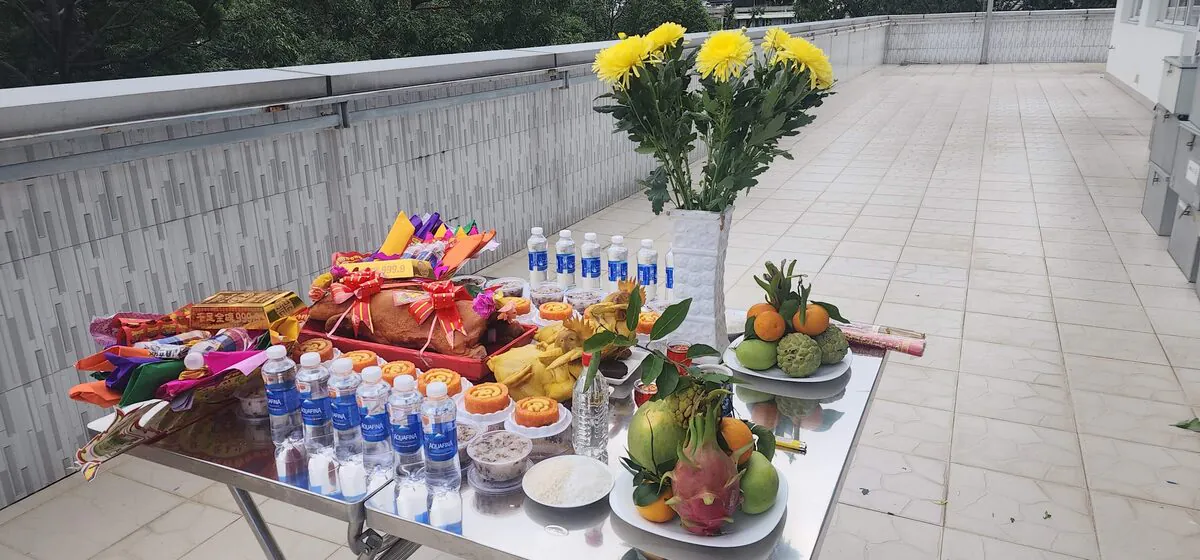 Góc chụp ngang mâm cúng cô hồn VIP trên sân thượng gồm heo quay nguyên con thắt nơ đỏ, gà luộc, bình hoa cúc vàng, 2 đĩa ngũ quả thanh long mãng cầu bưởi, xôi gấc, chè đậu, 12 chai Aquafina và quần áo chúng sinh
