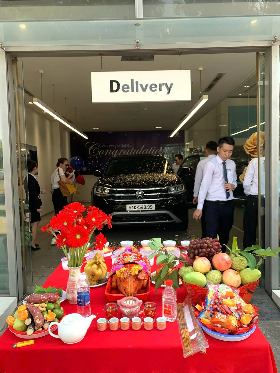 Mâm cúng xe mới đặt trước mũi xe SUV Volkswagen Teramont màu đen biển số 51K-543.99 tại phòng Delivery Showroom Volkswagen Sài Gòn gồm heo sữa quay, gà luộc, hoa đồng tiền đỏ và mâm ngũ quả