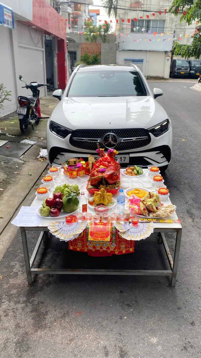 Mâm cúng xe mới đặt ngay trước mũi xe SUV Mercedes-Benz GLC màu trắng gồm heo sữa quay nguyên con, gà trống luộc, 6 đĩa xôi gấc chữ Lộc, bánh phu thê, ngũ quả và tiền giấy xếp hình quạt tại Chung Cư Đúng Nghi Thức — Ý nghĩa & nghi thức dâng lễ xe mới mua tại nhà chuẩn tín ngưỡng