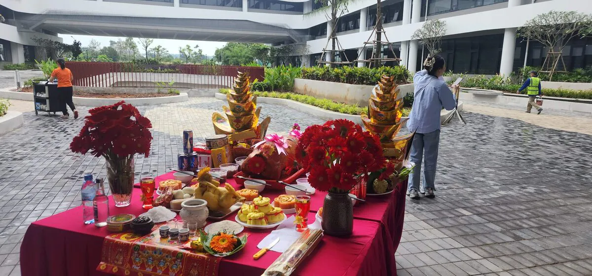 Toàn cảnh bàn đại lễ cúng Đất Đai tại sân nội khu tòa nhà thương mại với heo quay nguyên con, gà luộc, bánh tài lộc vàng, xôi gấc, 2 bình hoa đồng tiền đỏ và 2 tháp vàng