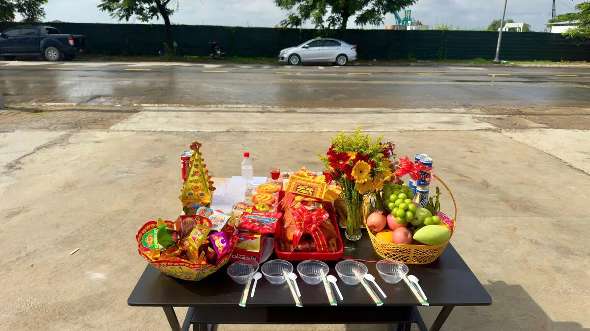 Mâm cúng động thổ đặt hướng ra mặt đường công trình gồm heo sữa quay thắt nơ đỏ, giỏ trái cây nho xanh táo đỏ xoài, bình hoa đồng tiền, tháp Kim Sơn và 5 bộ chén đũa