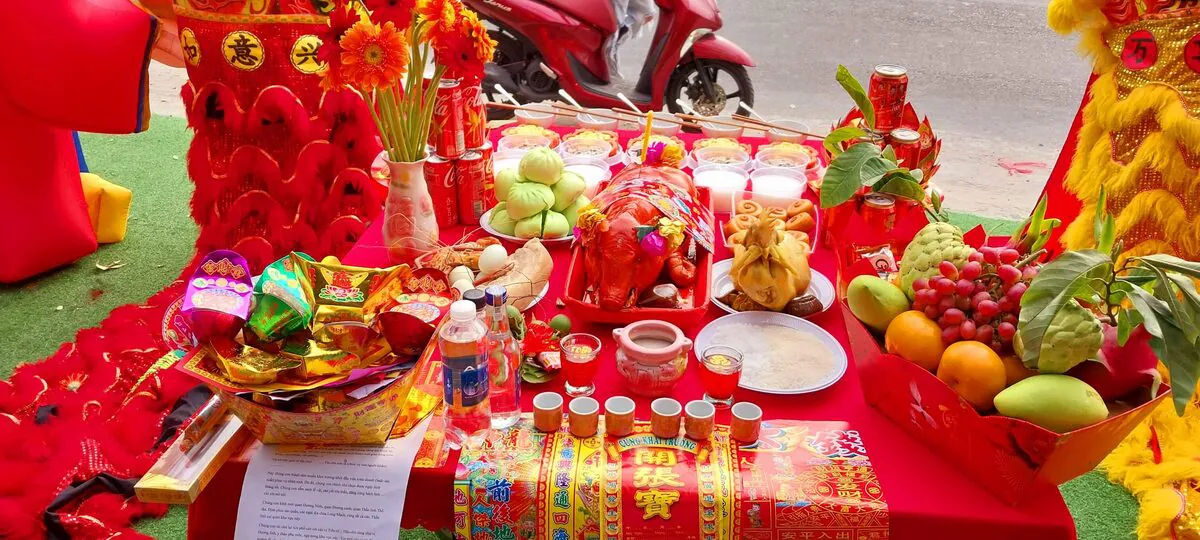 Cận cảnh mâm cúng khai trương giữa hai đầu lân đỏ vàng gồm heo quay sữa nguyên con, gà luộc, bộ tam sên, đĩa bánh bao lá dứa xanh, bánh bao chiên, khay trái cây nho đỏ mãng cầu xoài cam và tờ văn khấn khai trương