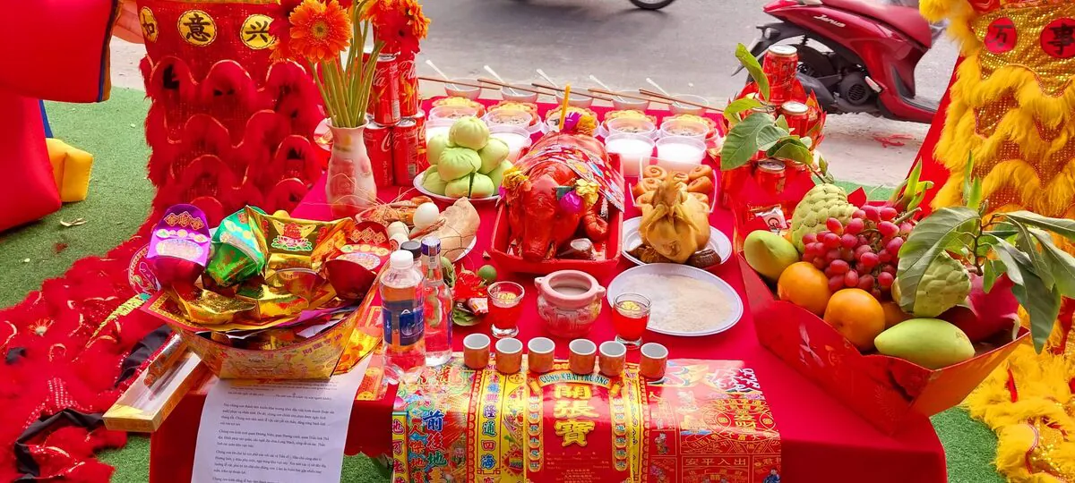 Mâm lễ cúng khai trương hồng phát đặt giữa cặp lân đỏ vàng gồm heo quay sữa, gà luộc, bộ tam sên, đĩa bánh bao xanh, khay trái cây tươi, mâm vàng mã và bộ ly trà rượu gạo muối