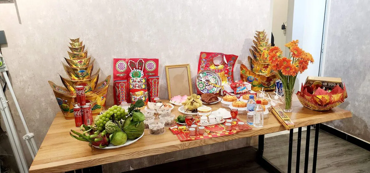 Mâm cúng Tổ Nghề trên bàn gỗ dài tại studio gồm bộ mã mão Quan Lớn hài đỏ, 2 tháp vàng, gà trống luộc, heo quay miếng, xôi gấc, chè đậu, nho xanh bưởi xanh và hoa đồng tiền cam